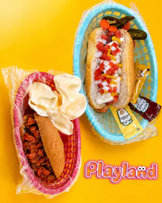 Un hot dog con tomate, cebolla y chiles y un chilidog con papas, servidos en canastas de plástico sobre fondo amarillo